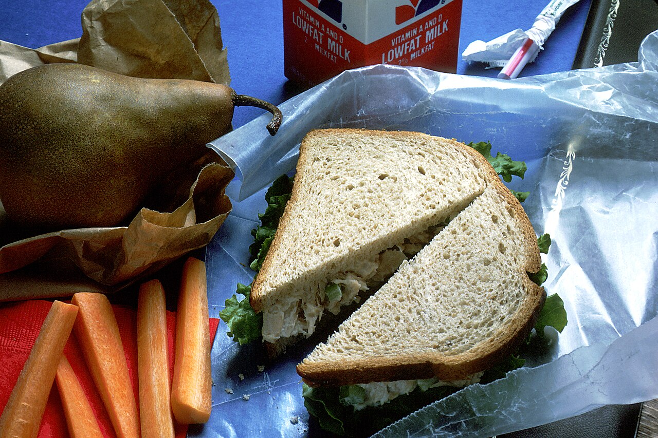 A packed brown-bag lunch with a sandwich, carrots, a pear, and a carton of milk