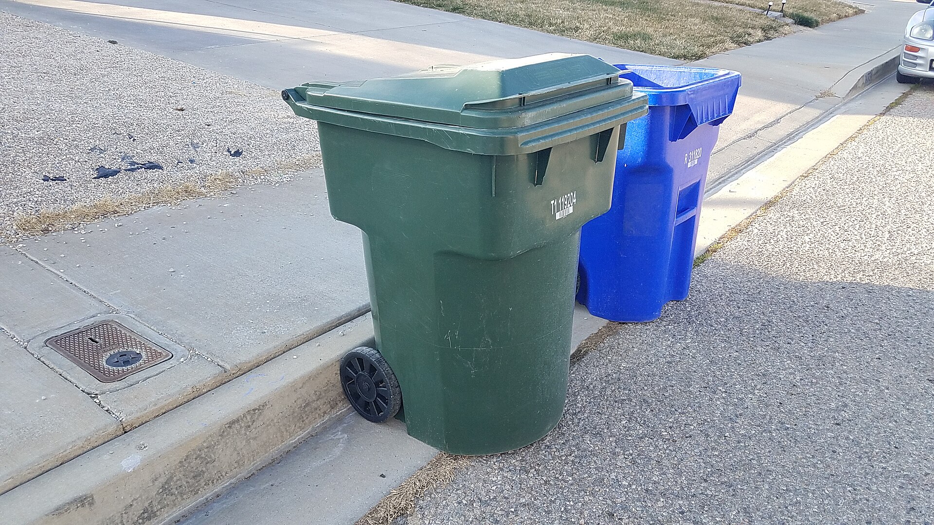 A green two-wheel trash cart at the street with one wheel pressed against the curb